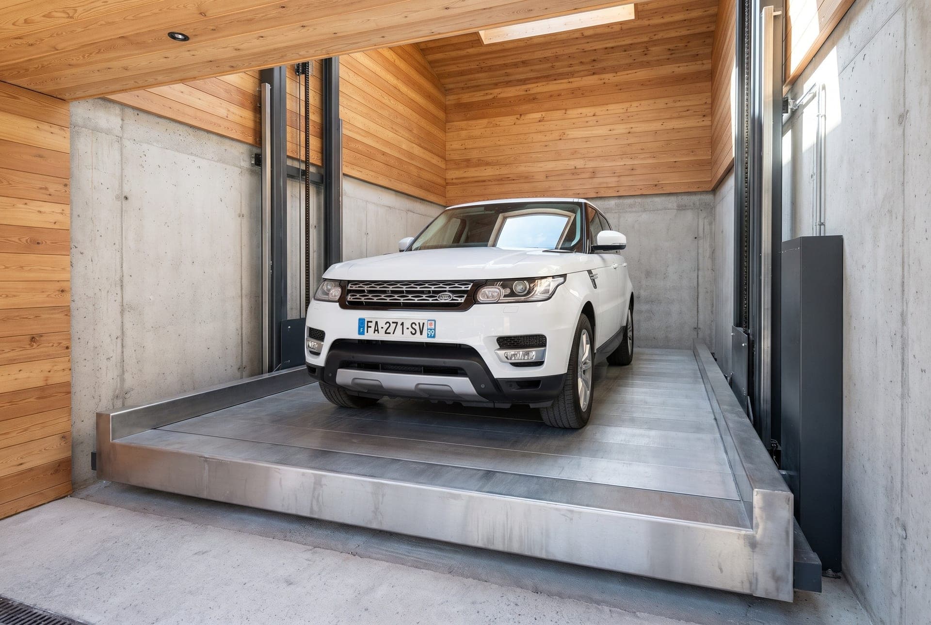 SUV de luxe blanc sur la plateforme acier d'un monte-voiture dans sa gaine béton, colonnes de guidage de part et d'autre, garage de chalet habillé de mélèze