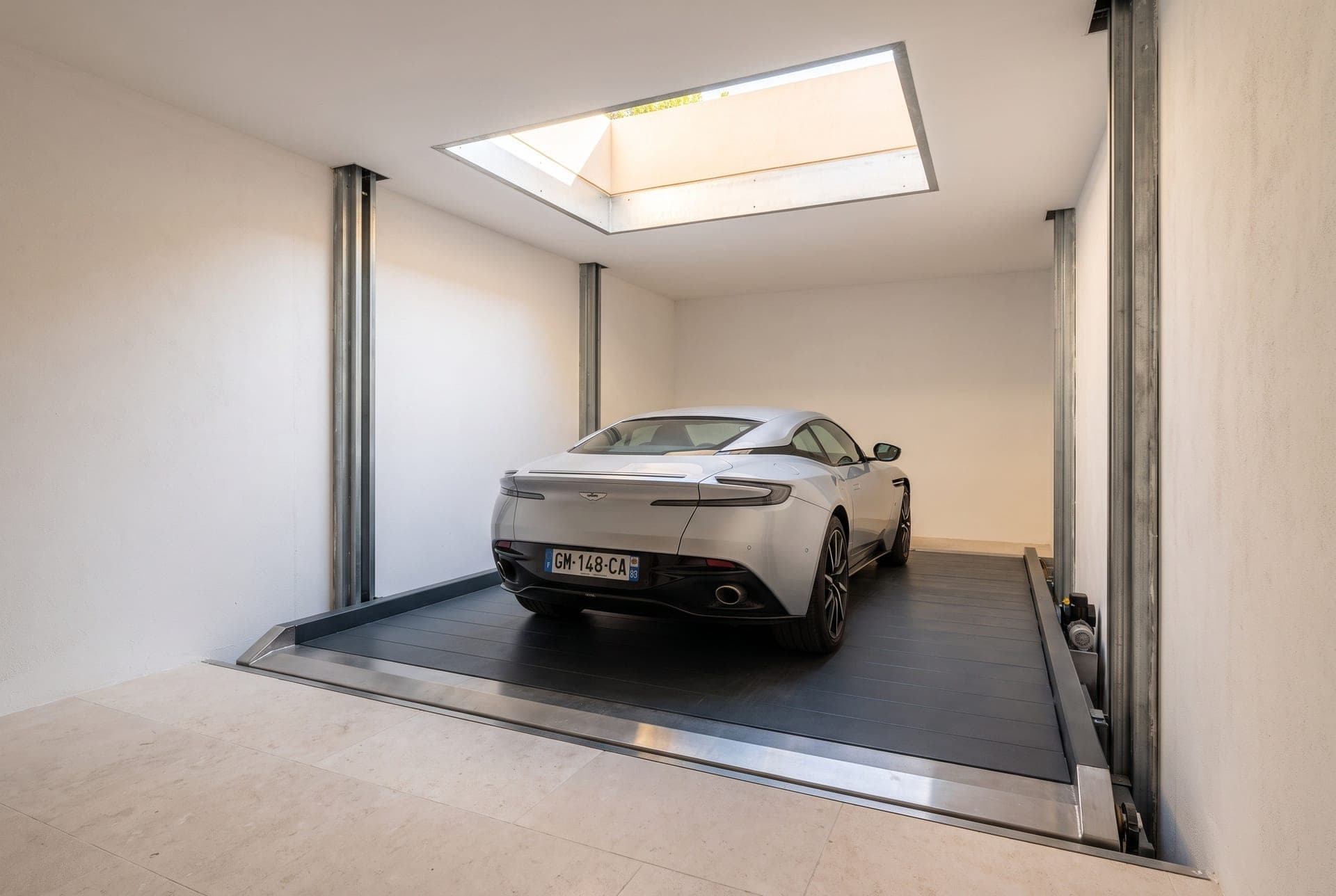 Coupé de luxe gris clair garé sur la plateforme d'un monte-voiture au fond de sa trémie béton blanche, rails de guidage le long des parois, lumière du jour tombant de la trappe