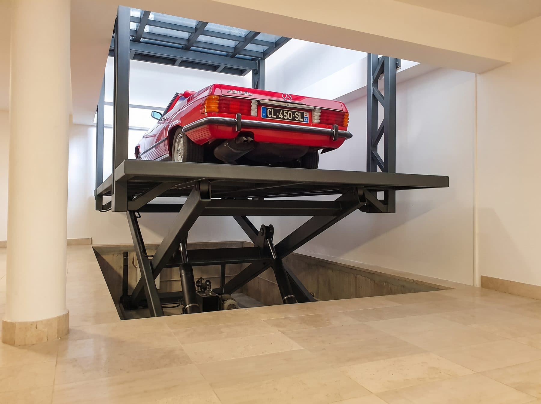 Monte-voiture à ciseaux : roadster rouge sur la plateforme, ciseaux et vérins déployés depuis la fosse en béton, cadre de guidage vers le niveau supérieur