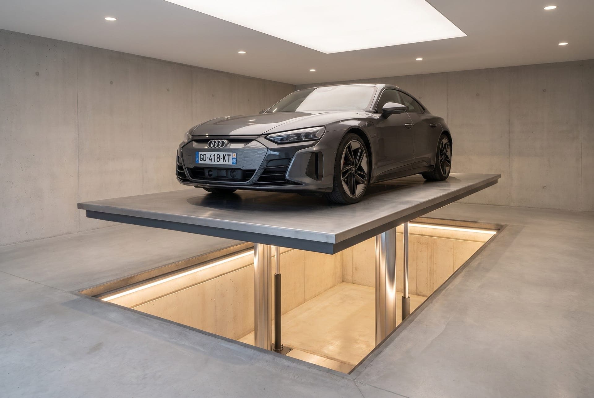 Monte-voiture hydraulique : coupé gris sur une plateforme unique au-dessus de la fosse éclairée, deux vérins chromés visibles dessous